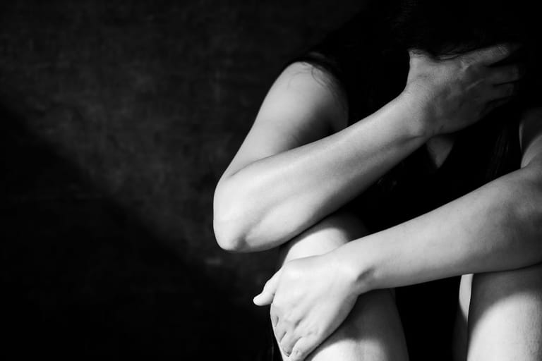 Kyyristyneenä istuva henkilö peittää kasvonsa käsillään tummassa ympäristössä, kuva välittää ahdistusta ja vaikeaa tunnetilaa.