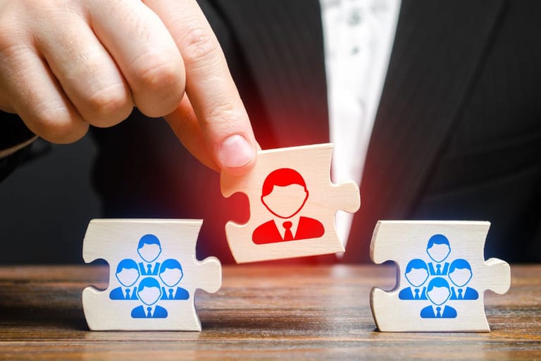 Käsi nostaa palapelin palaa, jossa on yhden henkilön kuvake, muiden palojen esittäessä ryhmiä ihmishahmoja pöydällä.