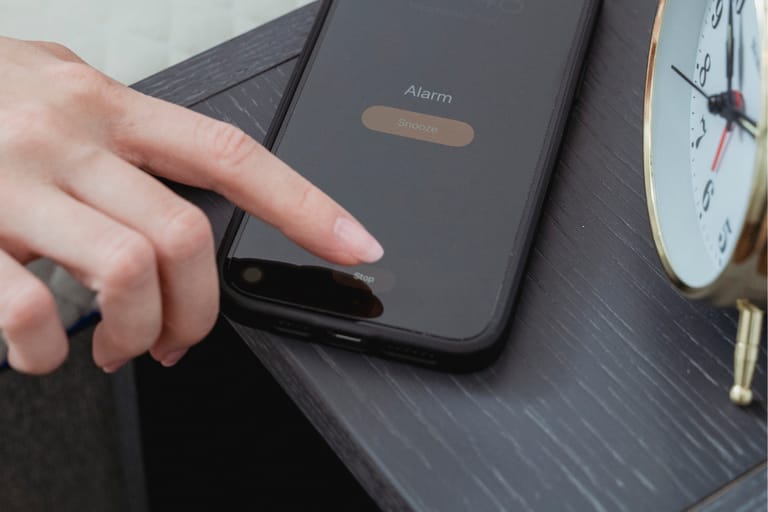 Sormi koskettaa älypuhelimen herätyskellon näytöllä olevaa “Stop”-painiketta yöpöydällä; vieressä näkyy perinteinen herätyskello.