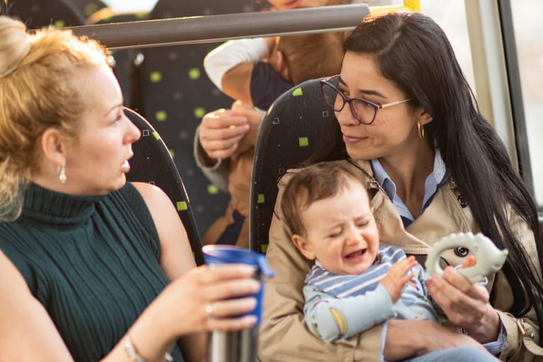 Kaksi naista istuu bussissa. Toinen nainen pitää sylissään itkevää vauvaa ja yrittää rauhoittaa häntä lelulla, samalla kun toinen nainen keskustelee hänen kanssaan ja pitää kädessään juomamukia. Taustalla näkyy muita matkustajia.