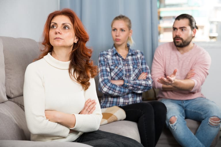 Kolme henkilöä istuu sohvalla sisätilassa. Edessä istuva henkilö valkoisessa neuleessa katsoo poispäin, kun taas taustalla oleva nainen ja mies keskustelevat, mies elehtii käsillään.