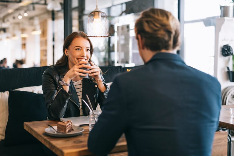 Nainen ja mies istuvat vastakkain kahvilassa; nainen hymyilee ja pitelee kuppia käsissään, pöydällä on kakku ja juomalasi, ja tunnelma on rento ja kiinnostunut.