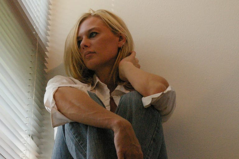 Vaalea hiuksinen henkilö istuu lattialla seinää vasten, polvet koukussa, katse sivulle suunnattuna ja ilme mietteliäs ikkunavalon osuessa kasvoihin.