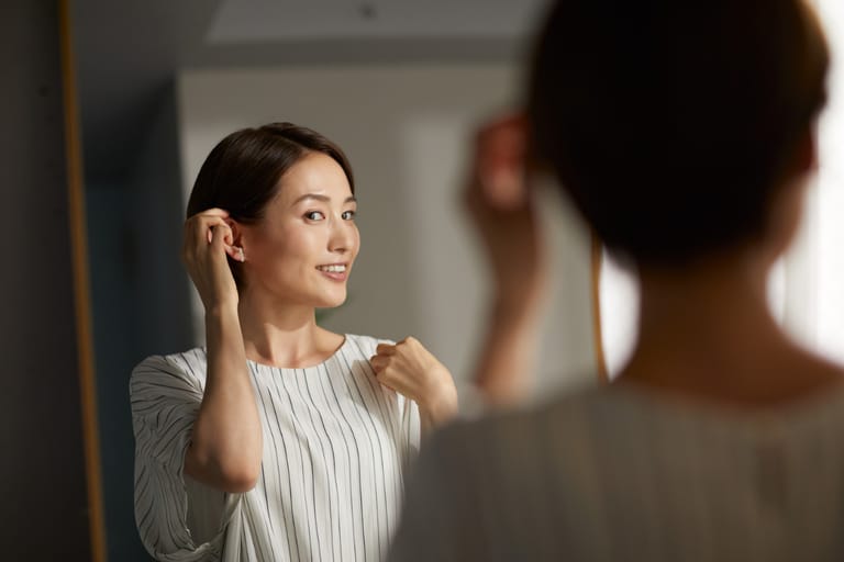 Henkilö katsoo peiliin ja koskettaa hiuksiaan, hänellä on vaalea pystyraidallinen paita ja rauhallinen, itsevarma ilme.