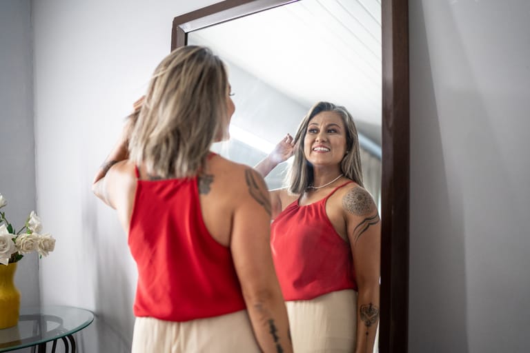 Nainen seisoo peilin edessä ja hymyilee omalle peilikuvalleen. Hänellä on punainen hihaton paita ja vaalea hame, ja hänen käsivarsissaan näkyy tatuointeja. Nainen kohottaa kättään hiustensa luo ja katsoo itseään itsevarmasti valoisassa huoneessa.