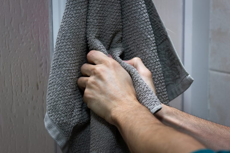 Lähikuva käsistä, jotka puristavat kosteaa harmaata pyyhettä kylpyhuoneessa; taustalla näkyy huuruinen suihkuseinä ja vesipisaroita.