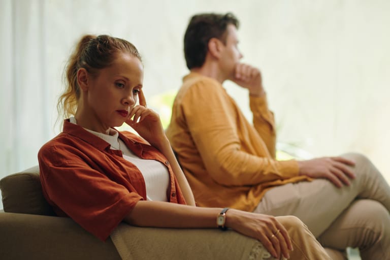 Nainen istuu sohvalla käsi poskea vasten ja katse alaspäin mietteliäänä, taustalla mies istuu selin häneen käsi suun edessä ja katse kaukaisuuteen, molemmat näyttävät etäisiltä ja huolestuneilta.