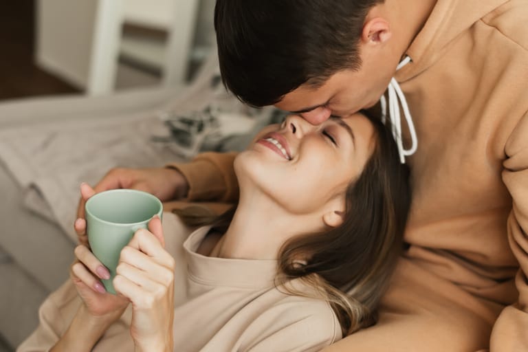 Nuori pari rentoutuu sohvalla kotona; mies suukottaa naista otsalle samalla kun nainen hymyilee silmät kiinni ja pitelee vihreää mukia käsissään, hetki huokuu läheisyyttä ja turvallisuutta.