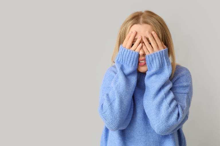 Nuori vaaleahiuksinen nainen sinisessä neuleessa seisoo vaaleaa taustaa vasten ja peittää kasvonsa molemmin käsin, ilmeestä päätellen nolostuneena tai vaivaantuneena.