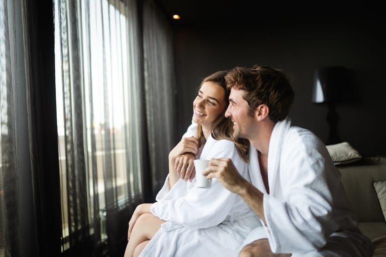 Kaksi henkilöä istuu vierekkäin lattialla vaaleissa vaatteissa ja pitää käsissään ruskeita kahvikuppeja; toisen käsi lepää toisen käsivarrella intiimissä ja rauhallisessa hetkessä.