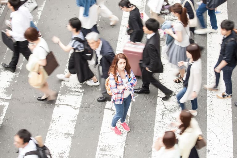 Nainen seisoo suojatiellä vilkkaassa kaupunkiympäristössä muiden ihmisten kulkiessa ohi.