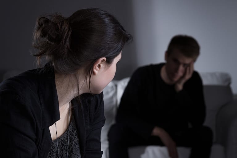 Nuori nainen istuu etualalla sisätiloissa ja katsoo sivulleen kohti miestä, joka istuu taustalla sohvalla pää painautuneena käteen. Tunnelma on etäinen ja kireä, ja molemmat vaikuttavat hiljaisilta ja huolestuneilta.