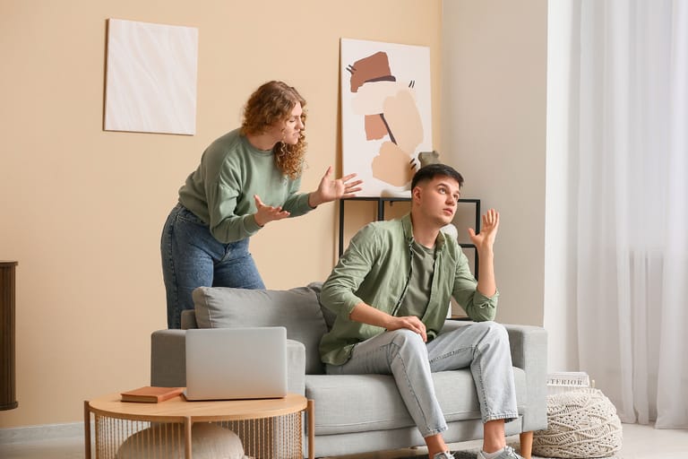 Nuori nainen seisoo olohuoneessa ja elehtii turhautuneena miehelle, joka istuu sohvalla ja näyttää välinpitämättömältä. Mies nojaa taaksepäin ja kohottaa kättään puolustautuvasti, kun nainen yrittää puhua. Tilanne kuvastaa ristiriitaa ja vallankäyttöä läheisessä ihmissuhteessa.