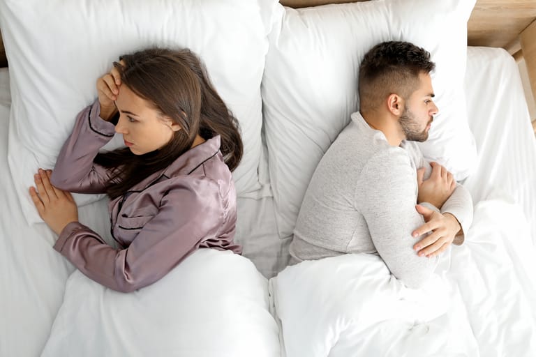 Nuori pari makaa sängyssä selät vastakkain. Nainen katsoo sivulle käsi otsallaan ja mies on kääntyneenä poispäin kädet ristissä, molemmat näyttävät etäisiltä ja mietteliäiltä. Valkoiset lakanat korostavat jännittynyttä ja etääntynyttä tunnelmaa.