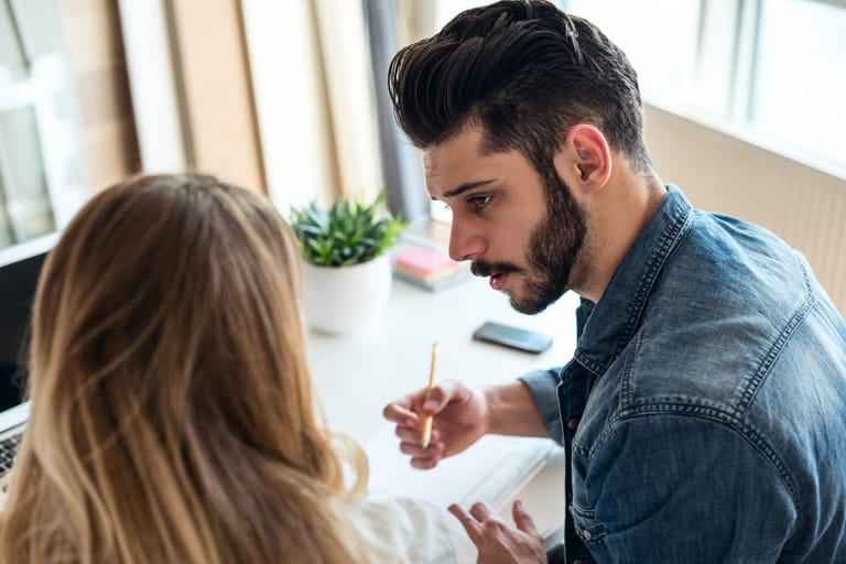 Nuori mies keskustelee keskittyneesti naisen kanssa työpöydän ääressä, pitää kynää kädessään ja katsoo naista vakavana, pöydällä on muistikirja, puhelin ja viherkasvi, tilanne viittaa neuvotteluun, ohjaukseen tai yhteiseen ongelmanratkaisuun.