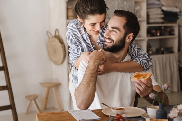 Nuori nainen halaa keittiössä istuvaa miestä takaapäin. Mies hymyilee ja pitää kädessään paahtoleipää, pöydällä on aamiaisastioita ja hilloa. Tunnelma on lämmin ja arkisen läheinen.