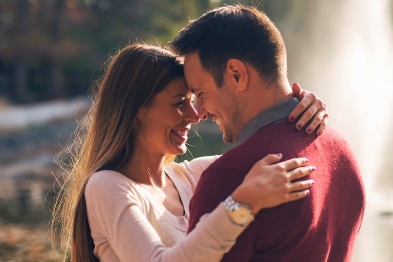 Hymyilevä pariskunta seisoo lähekkäin ulkona ja halaavat toisiaan otsat vastakkain lämpimässä auringonvalossa.