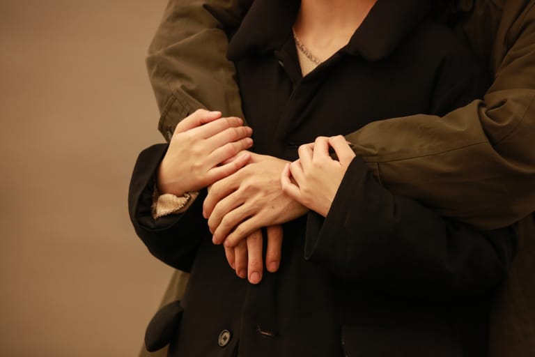 Kaksi ihmistä seisoo lähekkäin, toinen kietoo kätensä toisen ympärille takaapäin ja heidän kätensä lepäävät rinnakkain mustaa takkia vasten. Ele kuvastaa läheisyyttä, turvaa ja emotionaalista tukea.