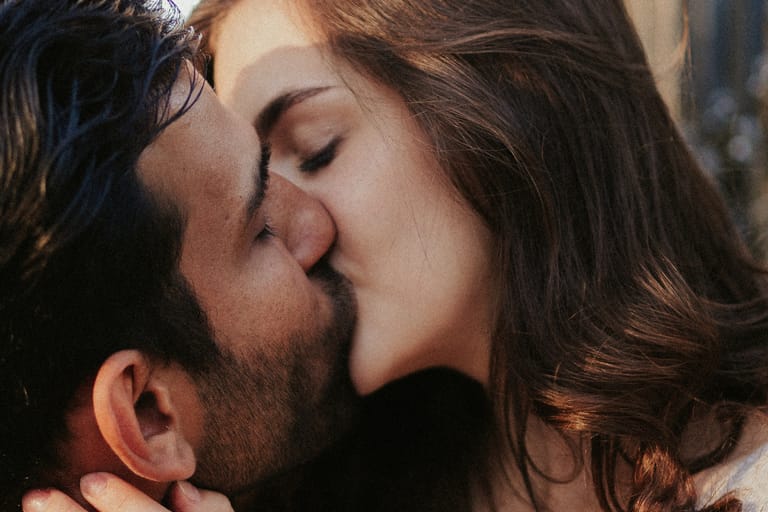 Lähikuva pariskunnasta suutelemassa toisiaan; mies ja nainen pitävät toisiaan kasvoista ja suutelevat intiimisti ulkona luonnonvalossa.