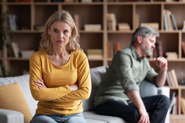 Olohuoneessa nainen istuu sohvalla kädet ristissä ja katsoo suoraan eteenpäin vakavana, taustalla mies istuu sivuttain katse poispäin, parin välillä vallitsee selvä riita ja emotionaalinen etäisyys.