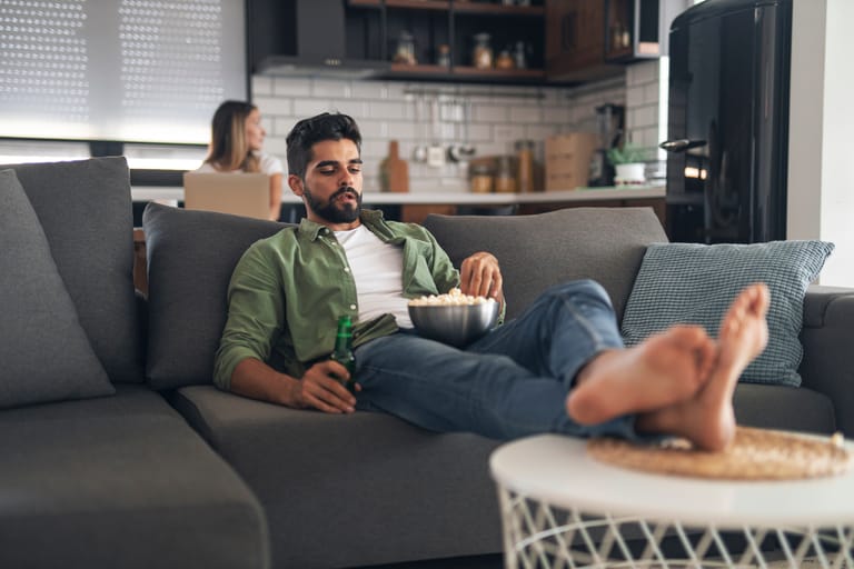 Mies makaa rennosti sohvalla jalat pöydällä, syö popcornia ja pitelee juomapulloa, kun nainen työskentelee kannettavan tietokoneen ääressä keittiössä taustalla.