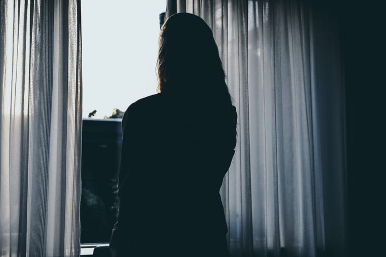 Siluettimainen hahmo seisoo ikkunan edessä puoliksi vedettyjen verhojen välissä ja katsoo ulos kirkkaaseen päivänvaloon. Tunnelma on hiljainen ja etäinen, ja vastavalo korostaa yksinäisyyden ja pohdinnan tunnetta.