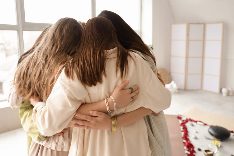 Kolme naista seisoo tiiviissä ryhmähalausessa valoisassa huoneessa, kädet toistensa selkien ympärillä. Tunnelma on lämmin ja lohduttava.