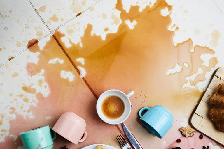 Lattialle ja pöydälle roiskunutta kahvia ja murusia; keskellä valkoinen kahvikuppi, ympärillä kaatuneita värikkäitä mukeja, kahvitahroja ja leivonnaisia.