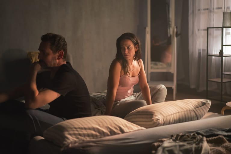 Pariskunta istuu sängyllä selät vastakkain hämärässä makuuhuoneessa; mies katsoo poispäin käsi kasvoilla, nainen istuu hiljaa ja katsoo häntä huolestuneena, tunnelma on etäinen ja jännittynyt.