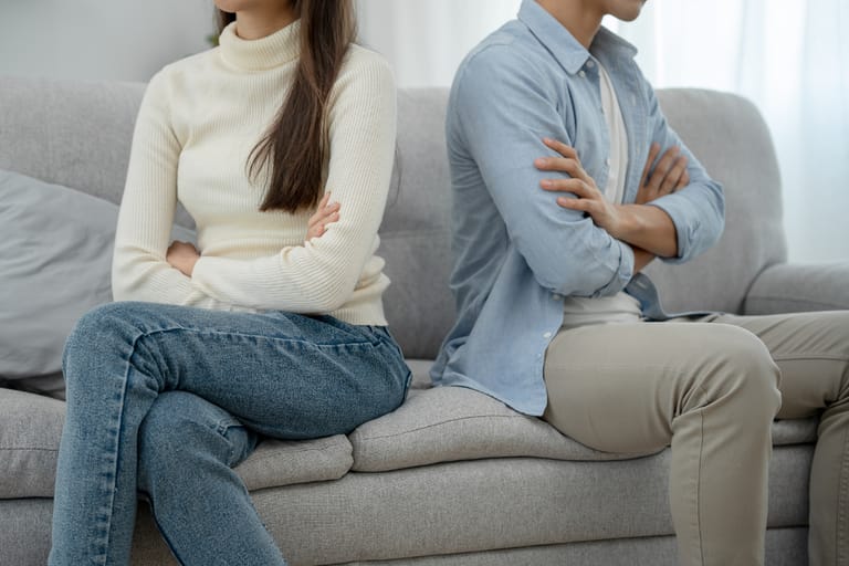 Nuori nainen ja mies istuvat sohvalla selät vastakkain ja kädet ristissä, ilmeeltään etäisinä ja jännittyneinä.