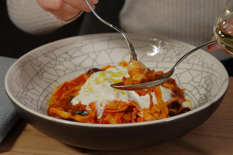 Haarukalla ja lusikalla nostetaan tomaattikastikkeessa olevaa pastaa, päällä burrataa tai pehmeää juustoa.