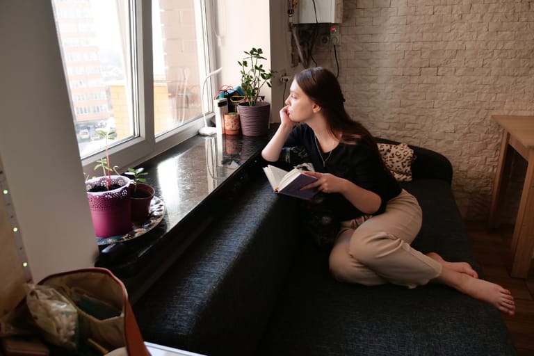 Nuori nainen istuu ikkunasyvennyksessä kotona, kirja kädessään. Hän nojaa päätään käteensä ja katsoo ulos ikkunasta mietteliäänä. Ikkunalaudalla on useita viherkasveja ruukuissa, ja huoneessa on rauhallinen, arkinen tunnelma.