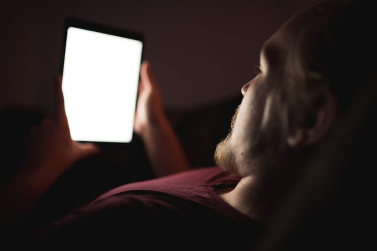 Mies makaa pimeässä ja katsoo kirkasta puhelimen näyttöä myöhään illalla.