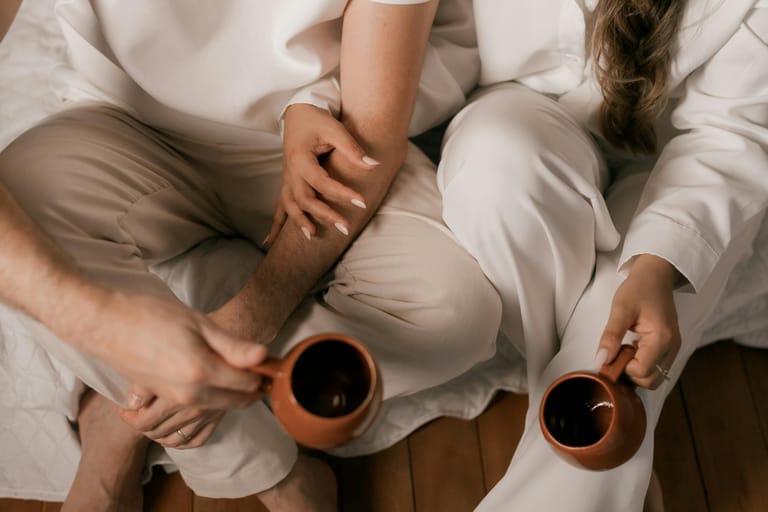Lähikuva kahdesta ihmisestä istumassa sängyllä vaaleissa vaatteissa; he pitelevät ruskeita kahvikuppeja ja toinen koskettaa lempeästi toisen käsivartta, mikä viestii rauhallisesta läheisyydestä ja yhteydestä.
