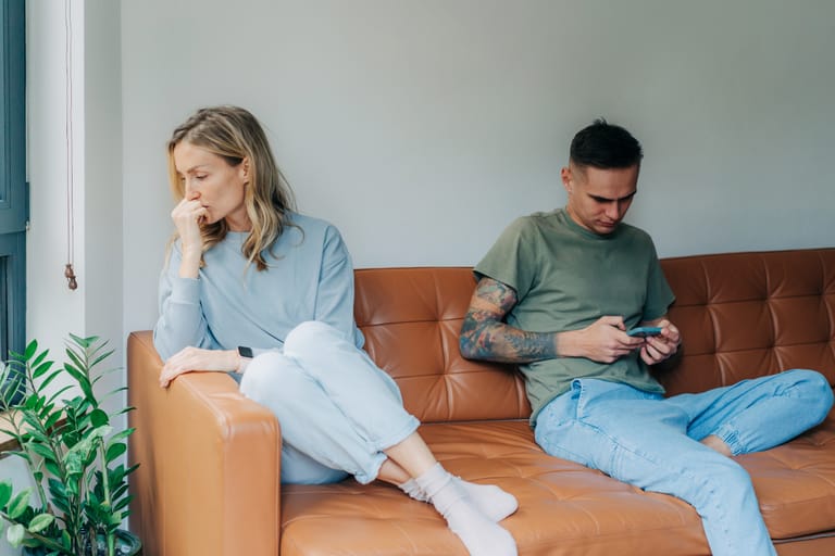 Nuori nainen istuu ruskealla sohvalla kädet koukussa ja katse poispäin, selvästi huolestuneena. Hänen vieressään mies istuu etäämmällä, katsoo puhelintaan eikä huomioi naista. Tunnelma on etäinen ja jännittynyt.