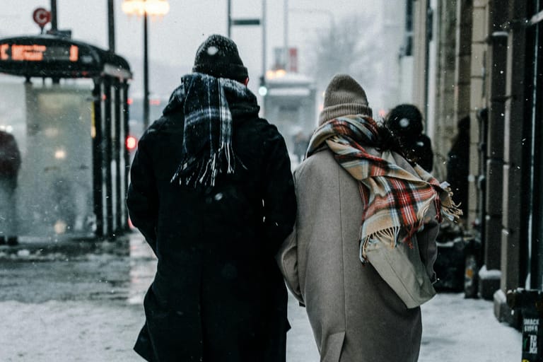 Kaksi talvivaatteisiin pukeutunutta ihmistä kävelee poispäin kamerasta lumisella kaupunkikadulla, lumisade ympärillään ja bussipysäkki taustalla.