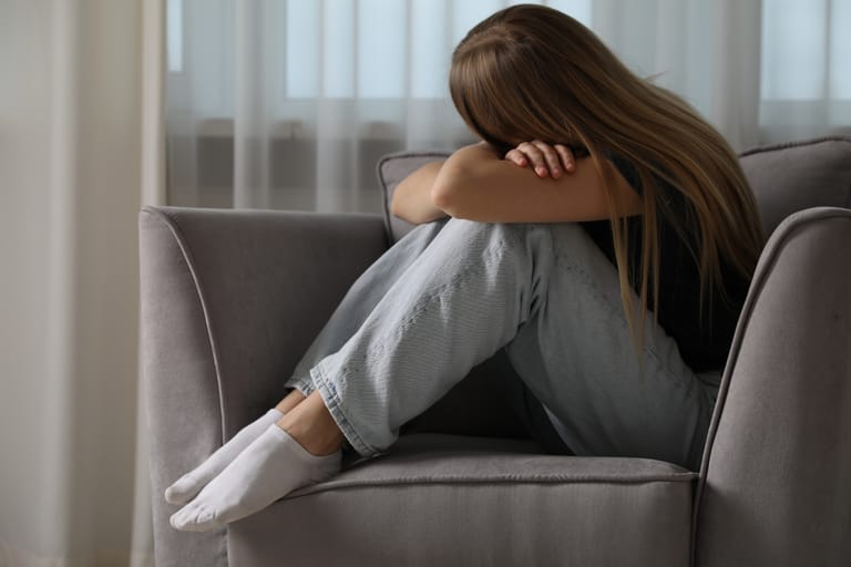 Nainen istuu sohvalla polvet rintaa vasten ja nojaa päätään käsiinsä, ilme kertoo ahdistuksesta.