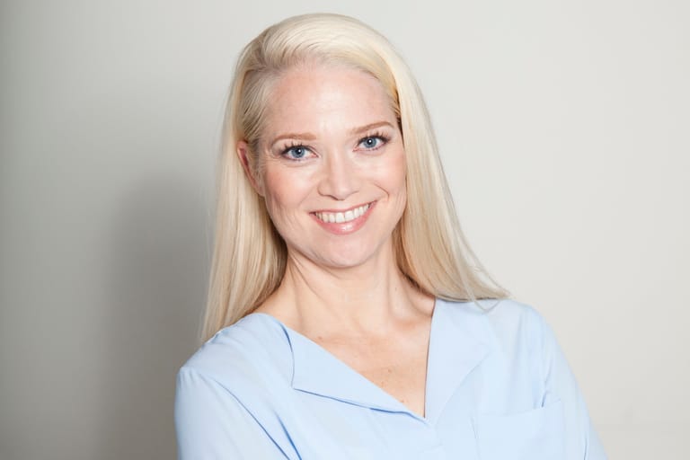 Pippa Laukka hymyilee valkoisen seinän edessä. Päällä vaaleansininen kauluspaita.