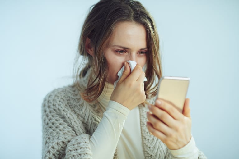 Henkilö pyyhkii nenäänsä paperinenäliinalla ja katsoo älypuhelimen näyttöä vaaleaa taustaa vasten.