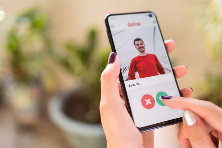 Käsi pitelee älypuhelinta, jossa on deittisovellus ja käyttäjä pyyhkäisee profiilia oikealle hyväksymisen merkiksi.