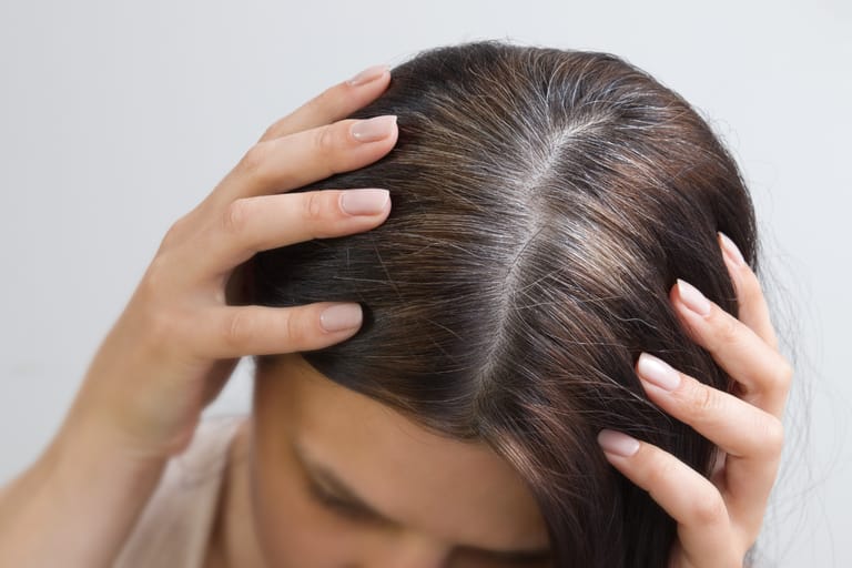 Lähikuva naisen päälaesta; hän pitelee hiuksiaan käsillään ja jakauksessa näkyy selvästi harmaita hiuksia tumman ruskean seassa.