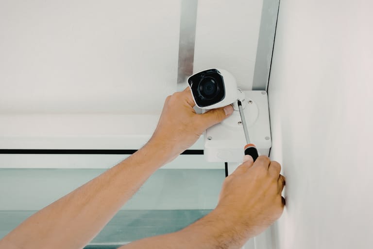 Henkilö asentaa valkoista valvontakameraa seinän yläkulmaan ruuvimeisselin avulla; kamera on kiinnitetty jakorasiaan katon rajassa.