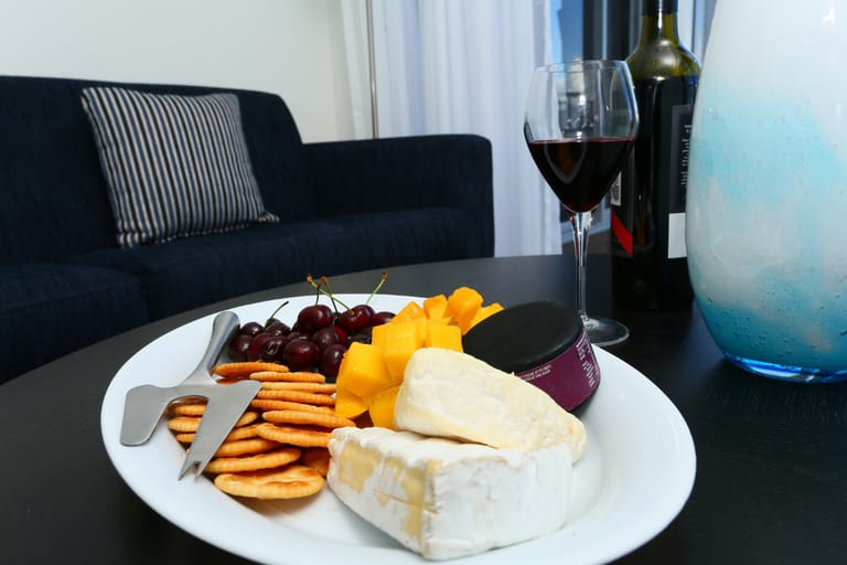 Juustolautanen pöydällä, jossa on briejuustoa, keksejä, viinirypäleitä ja hedelmiä, vieressä lasi punaviiniä ja viinipullo.