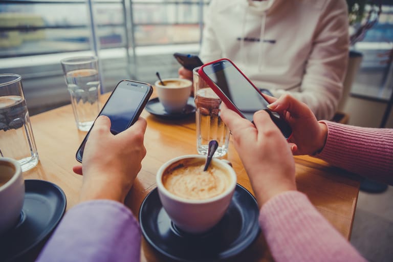 Usean henkilön kädet pitelevät älypuhelimia kahvilapöydän ääressä, pöydällä kahvikuppeja ja vesilaseja