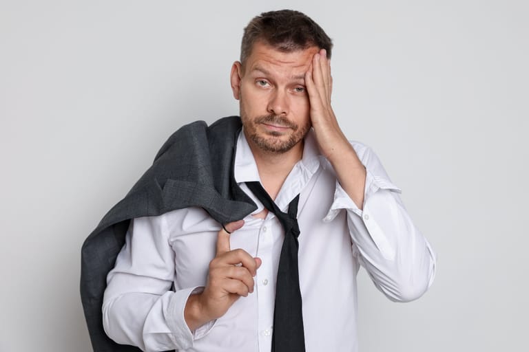 Mies valkoisessa kauluspaidassa ja löysällä solmiolla pitää kättään otsallaan väsyneen näköisenä, pikkutakki olkapäälle heitettynä vaaleaa taustaa vasten.