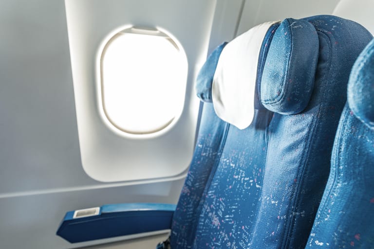 Lentokoneen ikkunapaikka: tyhjä sininen istuin ja kirkas ikkuna, josta tulvii valoa.