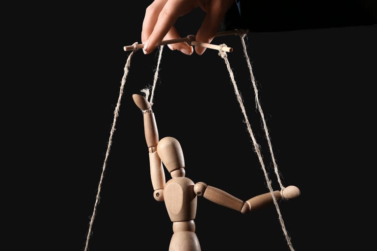 Puinen mallinukke roikkuu naruista kuin marionetti, ja käsi pitelee ohjausristikkoa mustaa taustaa vasten.