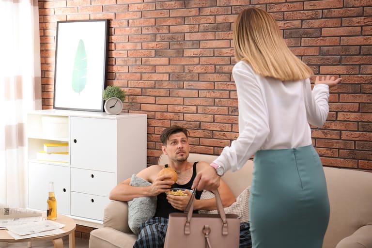 Nainen seisoo olohuoneessa käsilaukku kädessään ja elehtii miehelle, joka istuu sohvalla syöden hampurilaista ja katsoo häntä yllättyneenä.