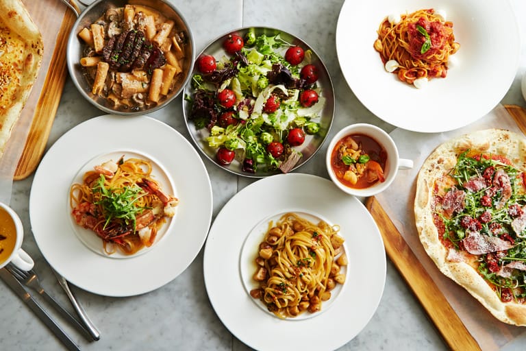 Ylhäältä kuvattu italialaistyylinen pöytä katettuna useilla annoksilla: pastoja, salaatti kirsikkatomaateilla, pizza, keitto ja leipää vaalealla marmoripöydällä.