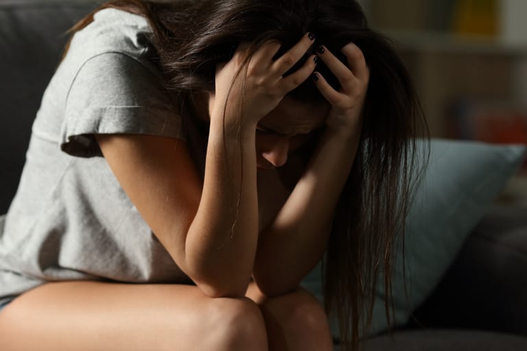 Nainen istuu etukumarassa ja pitelee päätään käsissään, ilme surullinen ja ahdistunut.